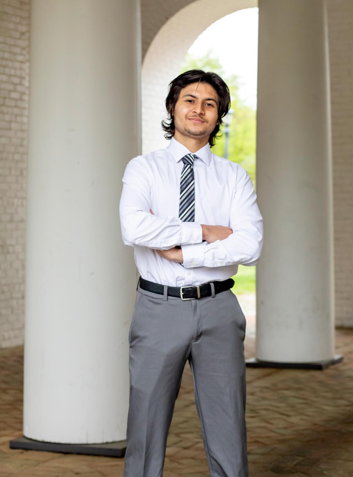 Male student dressed in a suit stands in a historical breezeway with arms crossed