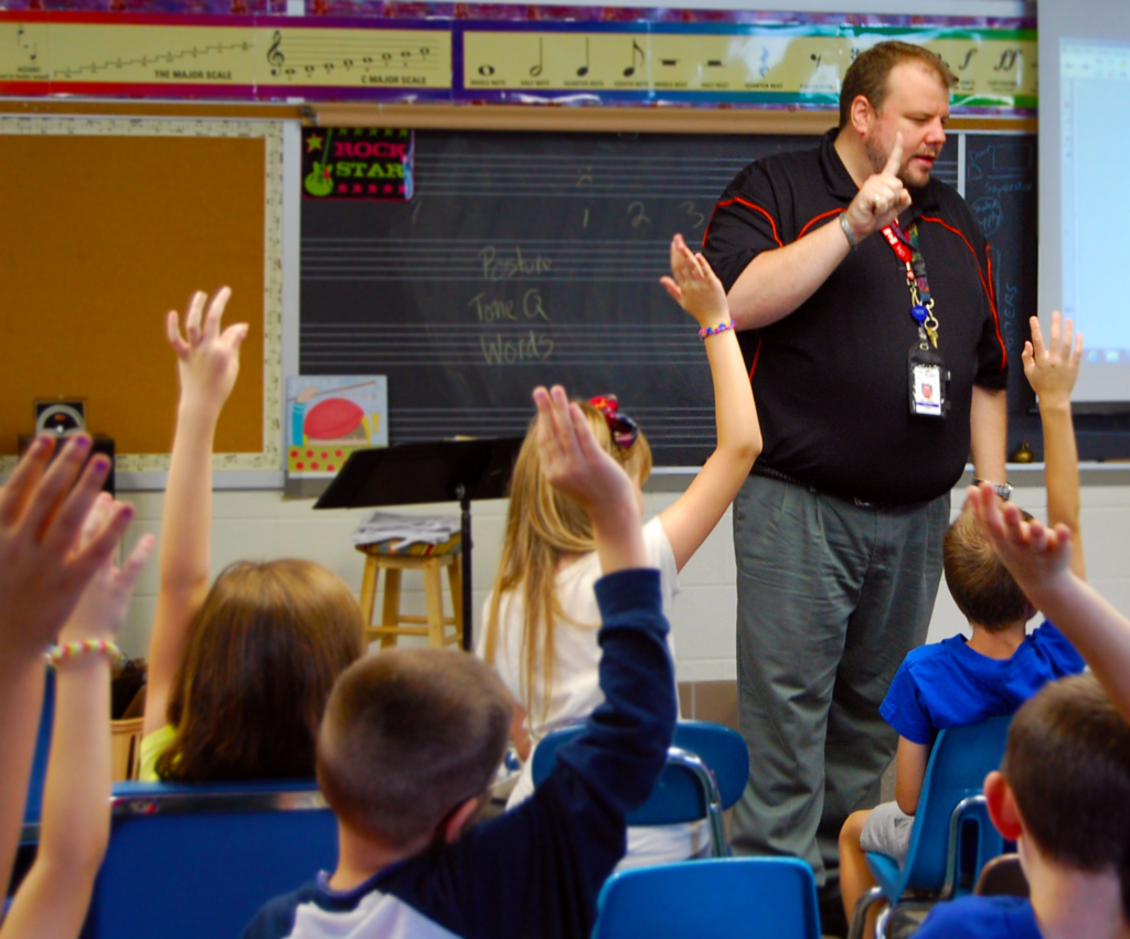 A student teacher asks the class a question and many students raise their hands.