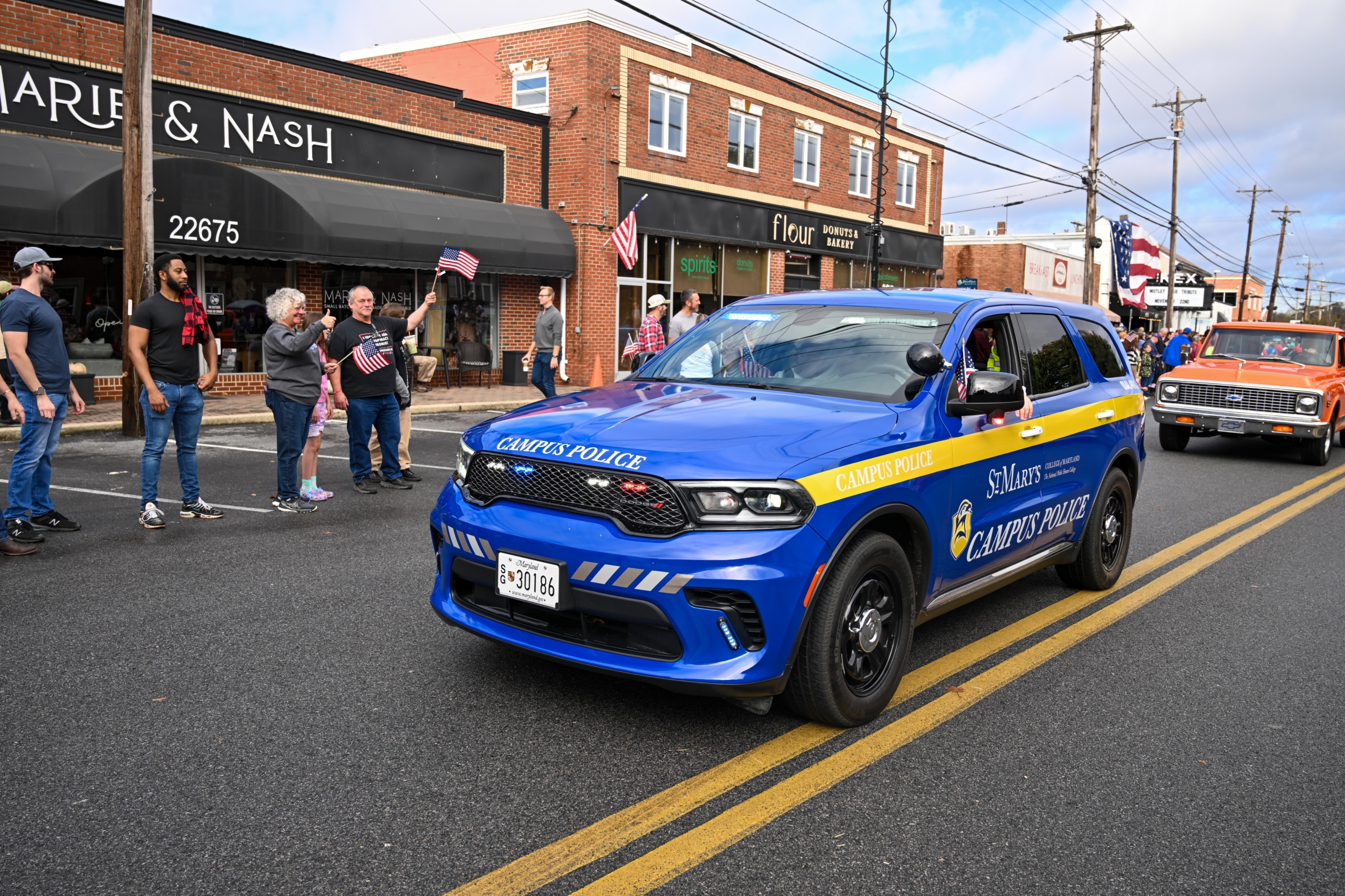A blue St. Mary’s Campus Police SUV drives down a street during a parade, passing spectators and other vehicles in front of shops.