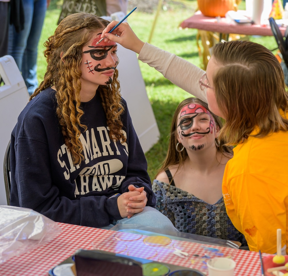 Photo by Chuck Steenburgh A person paints a pirate design on a girl's face while another girl with similar face paint looks on at an outdoor event.