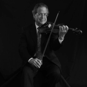 A man in a suit and tie is seated and playing a violin against a dark, plain background.