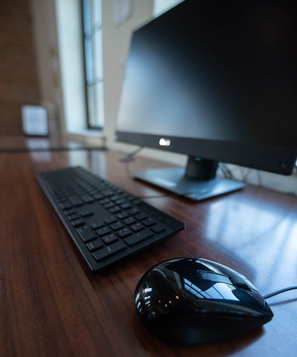 A computer monitor, keyboard, and mouse are arranged on a wooden desk near a window.