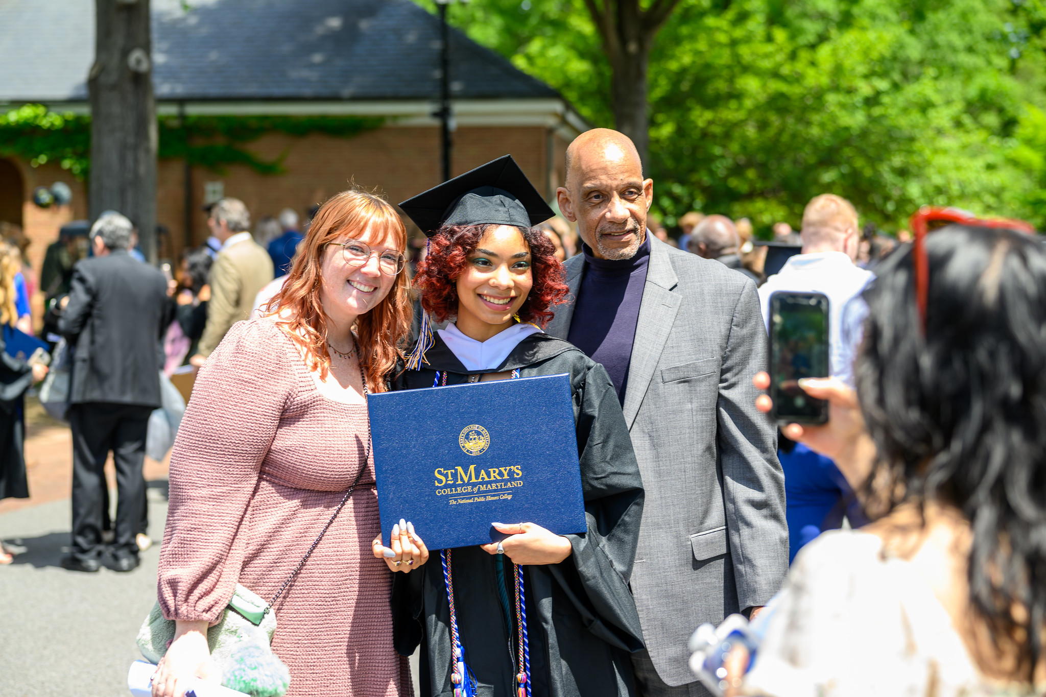 A graduate in cap and gown holds a diploma from St. Mary's College while posing with two people for a photo at an outdoor graduation ceremony.