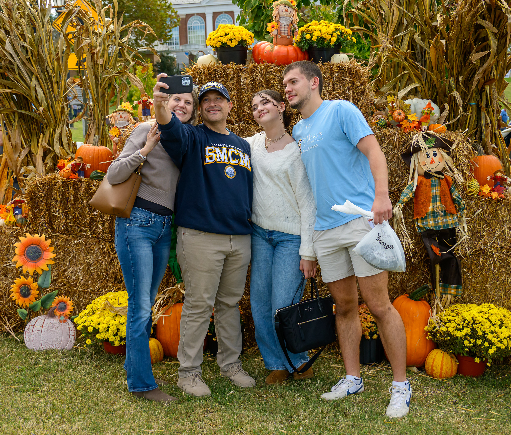 Four people stand in front of autumn decorations with pumpkins and hay bales, taking a group selfie outdoors.