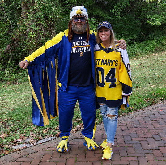 Two people pose outdoors; one wears a blue and yellow eagle mascot costume, and the other wears a yellow St. Mary's sports jersey and cap.