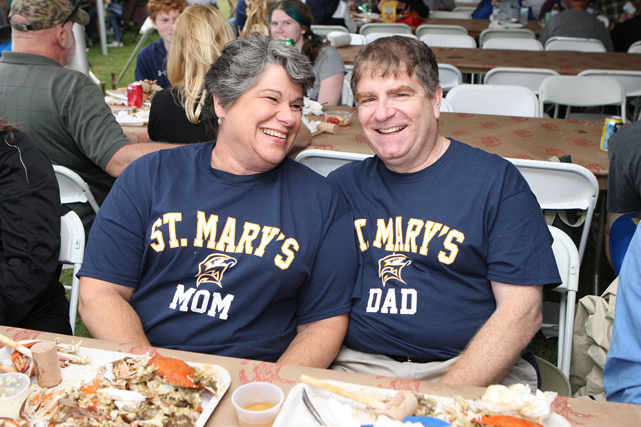 Two adults wearing "St. Mary's Mom" and "St. Mary's Dad" shirts sit and smile at a picnic table covered with food, surrounded by other people at an outdoor event.