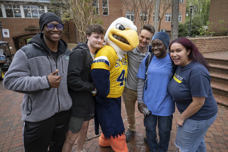 A group of five students poses outdoors with a person in an eagle mascot costume, all smiling for the camera.