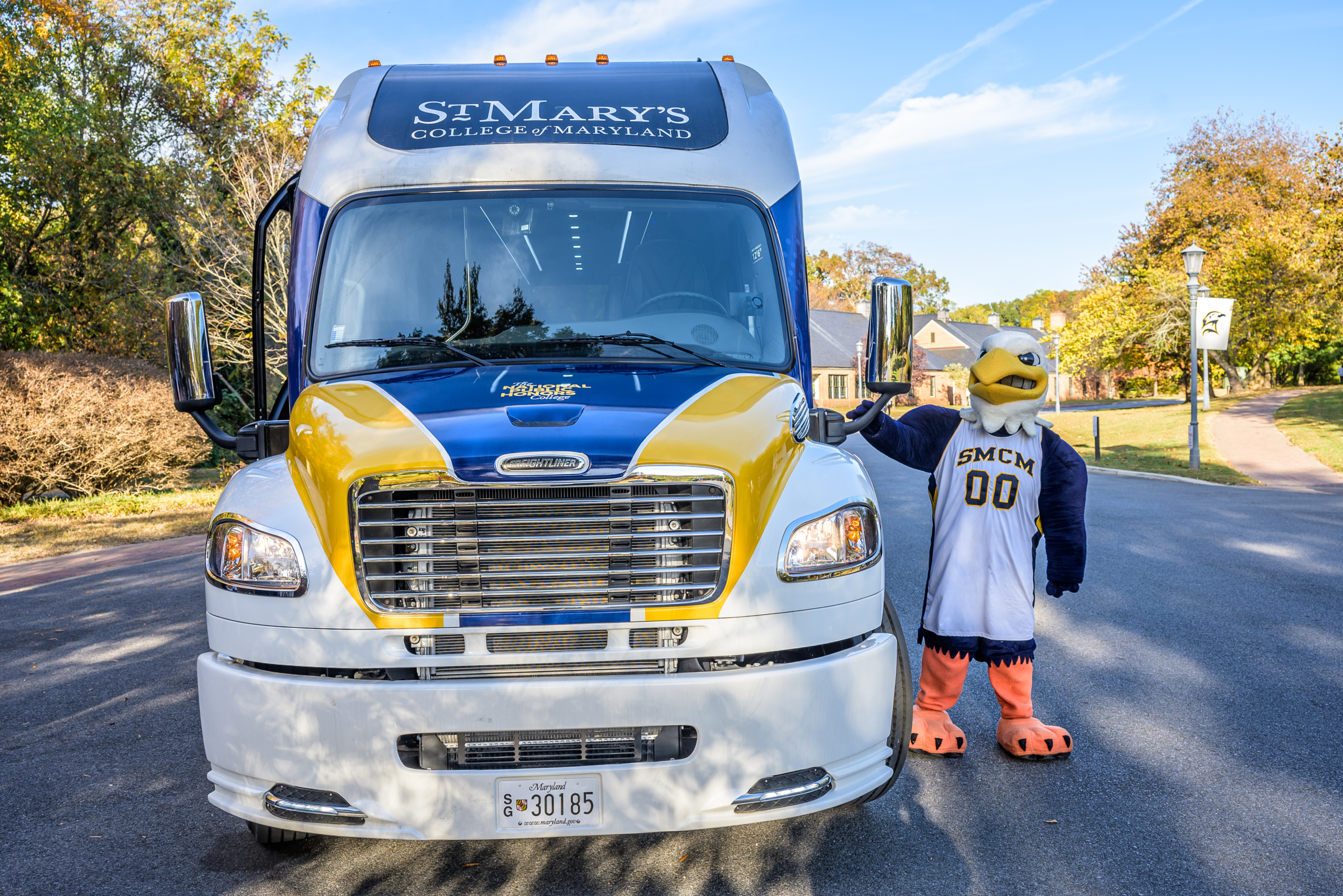 A person in a St. Mary's College of Maryland mascot costume stands beside a branded bus parked on a sunny road.