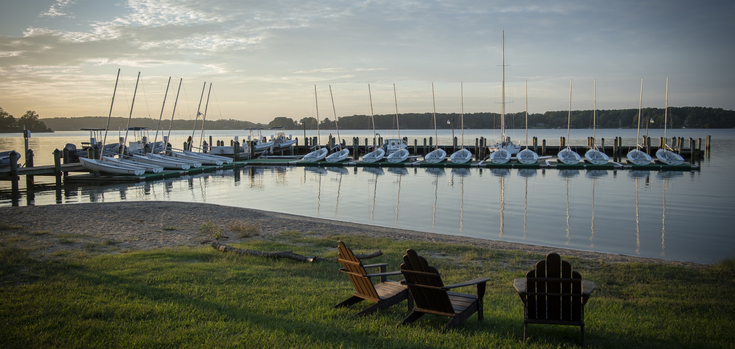 Two wooden chairs, reserved for Honors College students, face a calm body of water with rows of sailboats docked at the St. Mary's College of Maryland Waterfront
