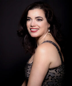 Woman with dark hair and red lipstick wearing a patterned sleeveless dress and dangling earrings, smiling against a dark background.