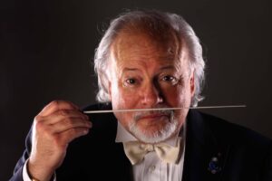 An older man with white hair and beard, wearing a tuxedo and bow tie, holds a conductor's baton in front of his face against a dark background.