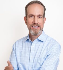 Middle-aged man with short brown hair and beard, wearing a light blue checkered dress shirt, standing with arms crossed and smiling at the camera against a plain background.