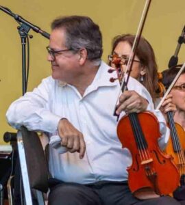 A man in a white shirt sits holding a violin and bow, with other musicians and microphones visible in the background.