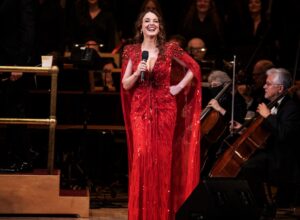A woman in a red, sequined gown stands on stage holding a microphone, smiling, with musicians seated behind her playing string instruments.