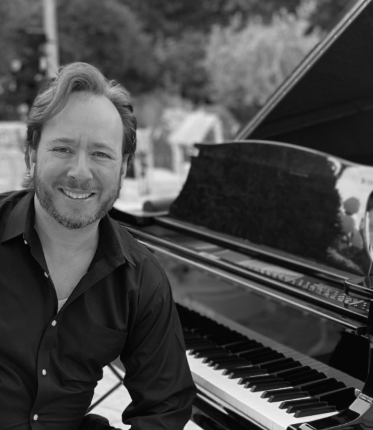 A man with a beard and wavy hair sits beside a grand piano outdoors, smiling at the camera. The image is in black and white.