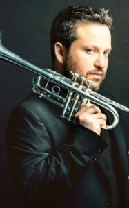 A man in a dark suit holds a trumpet resting on his shoulder, looking directly at the camera against a dark background. Instagram interface elements are visible on the right.