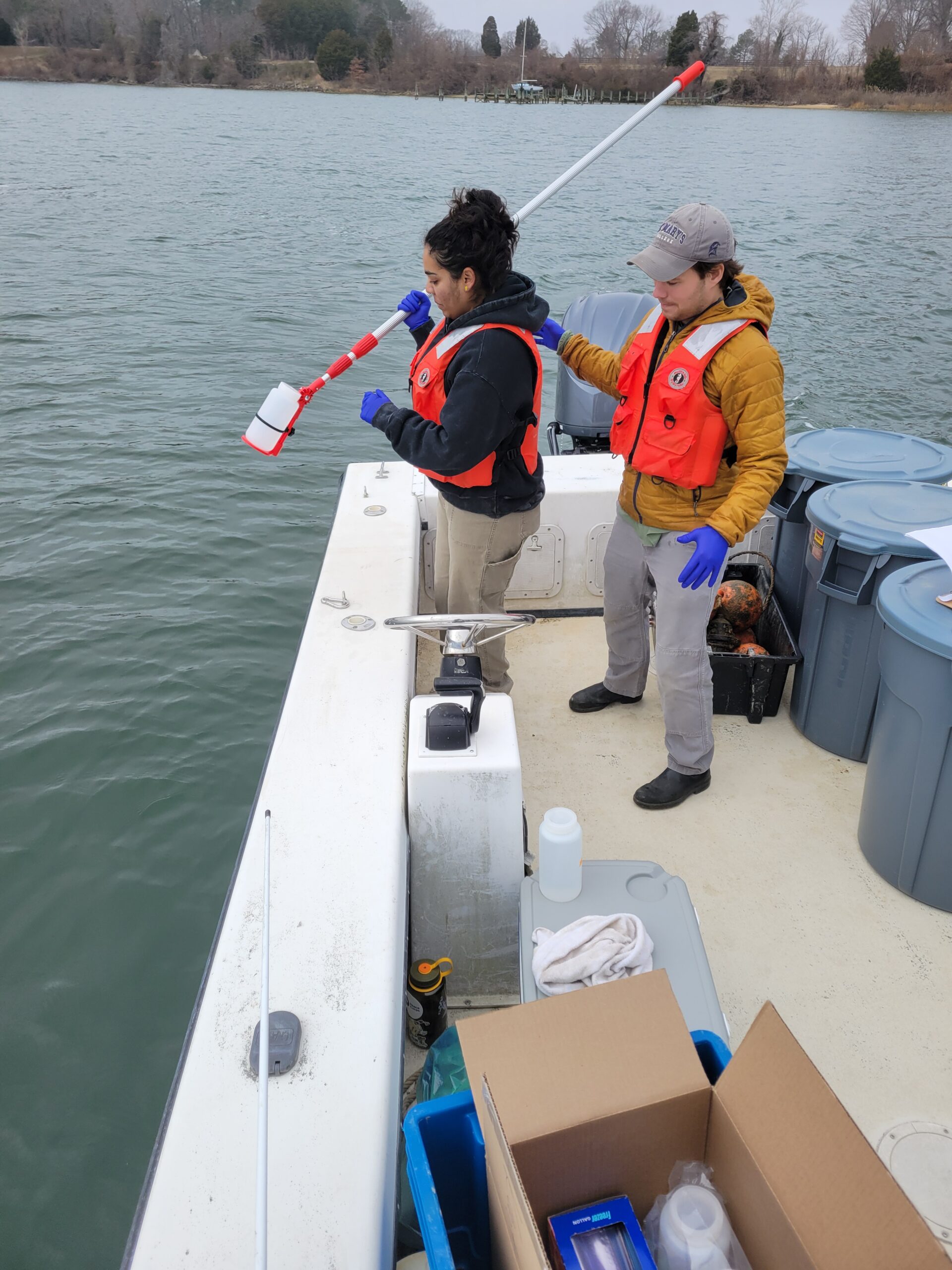 Two people in safety vests collect a water sample from a lake while standing on a boat equipped with scientific gear and storage bins.