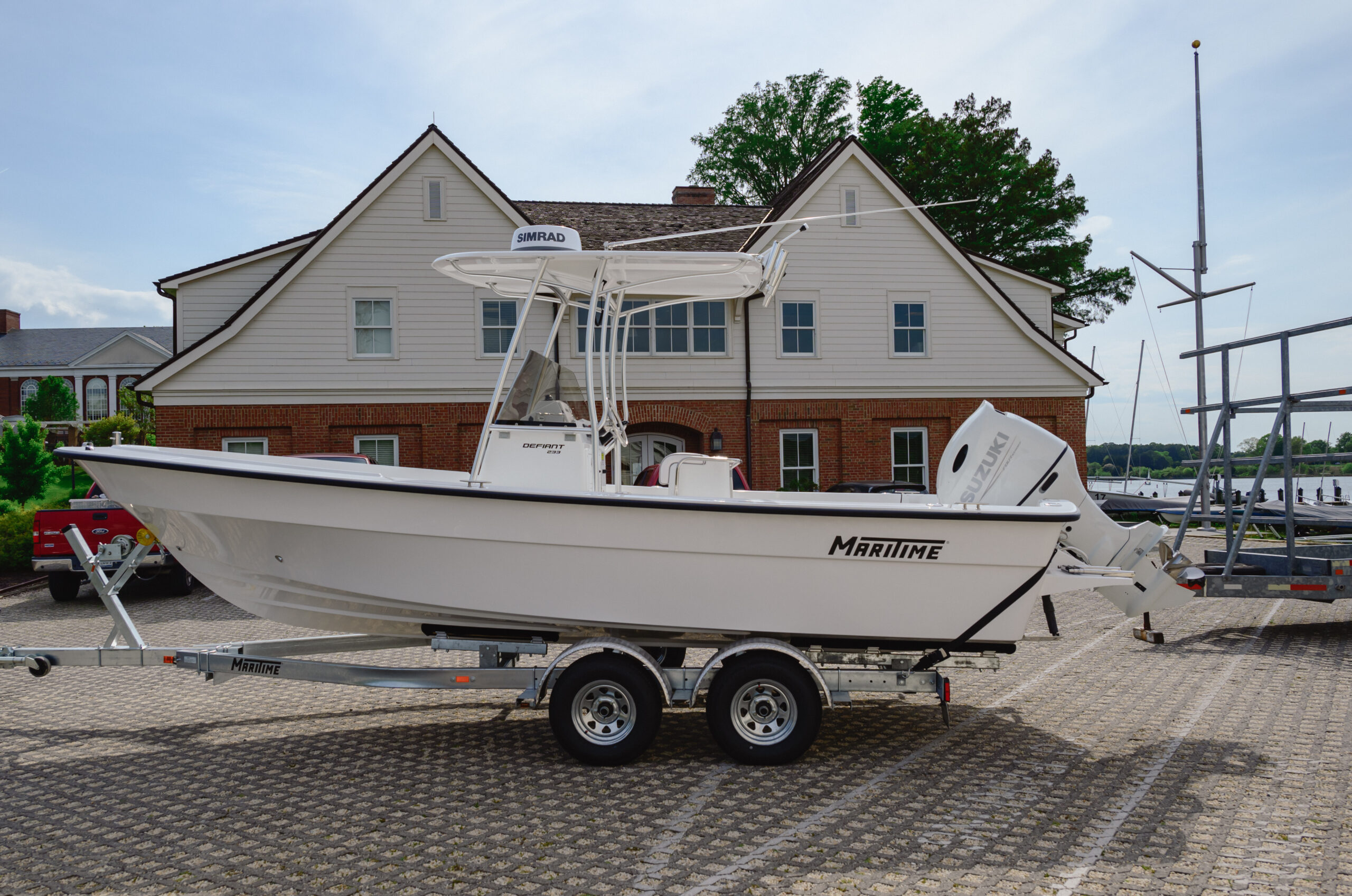 A white Maritime boat on a trailer is parked on a paved lot in front of a brick and white house, with trees and other boats in the background.