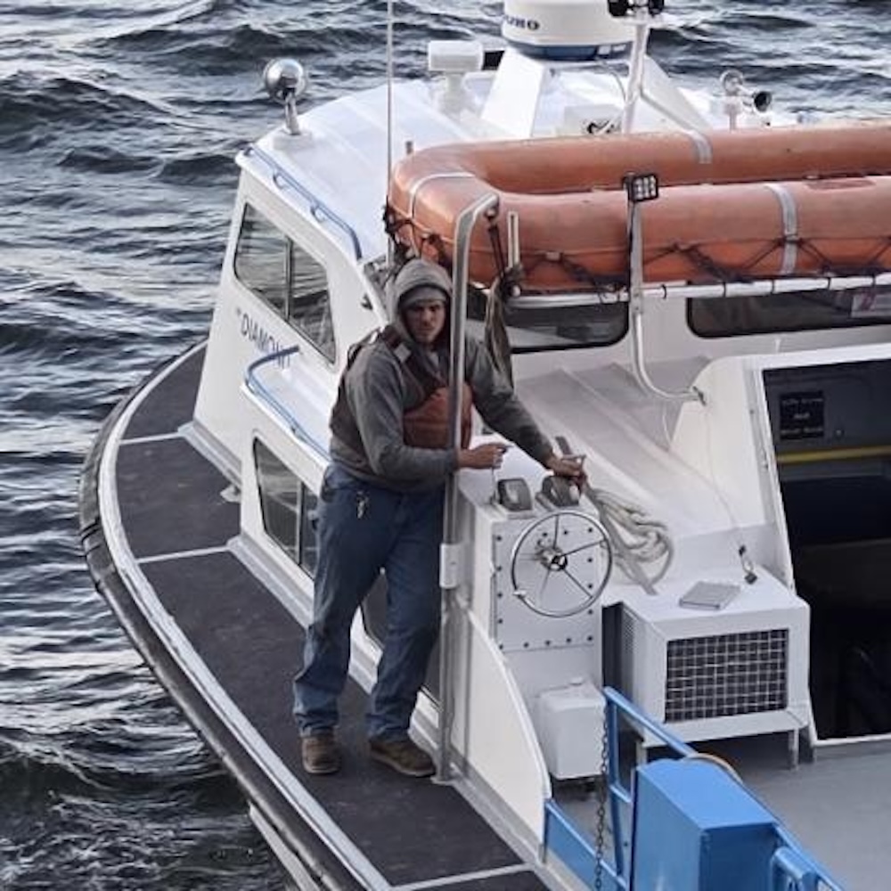 Person in work clothes stands on the deck of a small boat near the steering wheel, with life rafts stored on the roof and choppy water in the background.