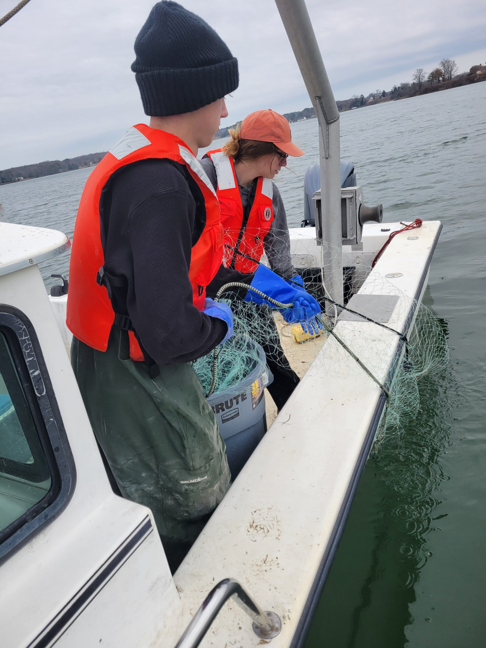 Two people in orange life jackets pull a fishing net out of the water while standing on a boat.
