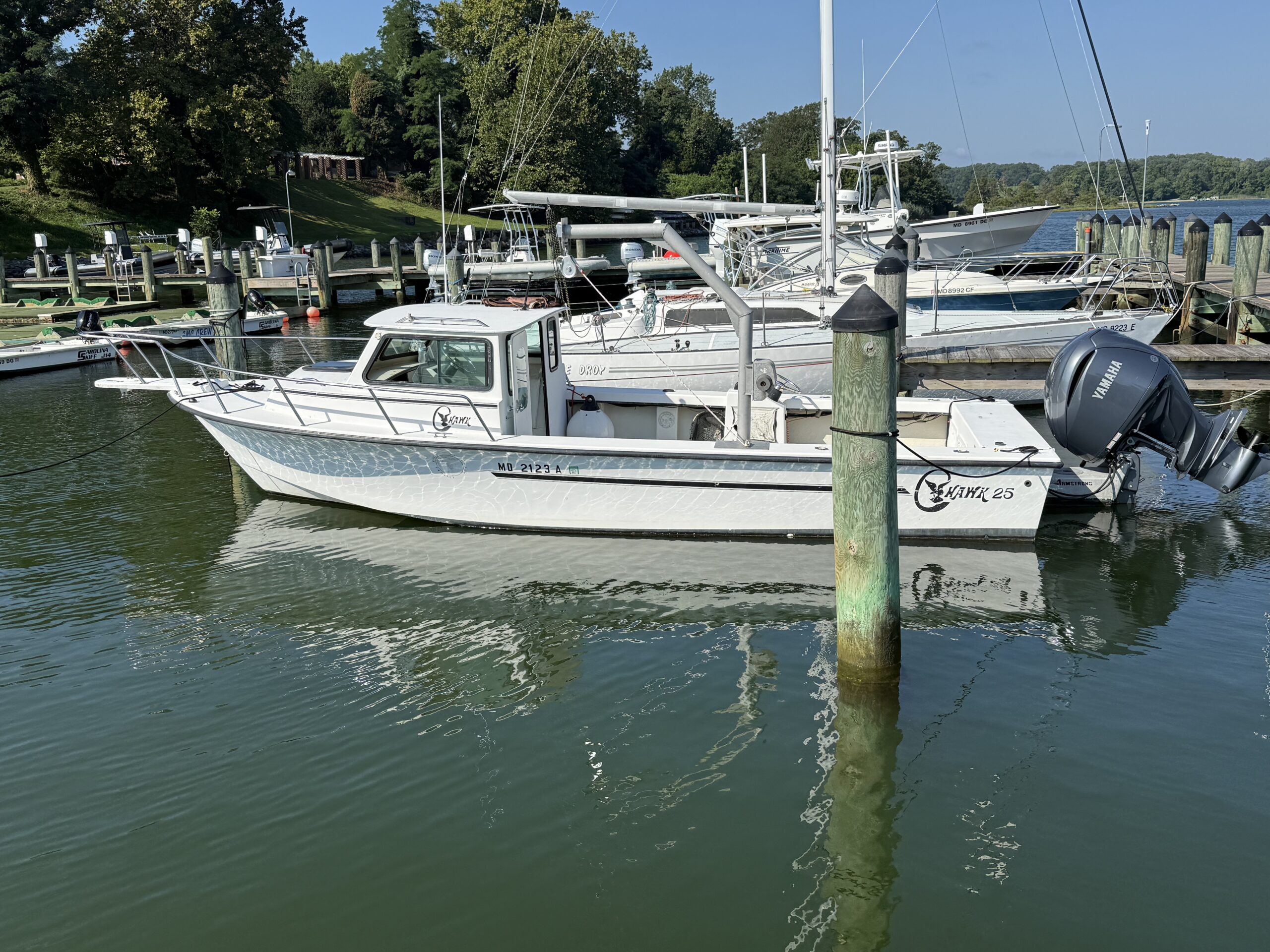 White fishing boat docked at a marina with outboard motors, other boats, green trees, and wooden pilings visible in the background.