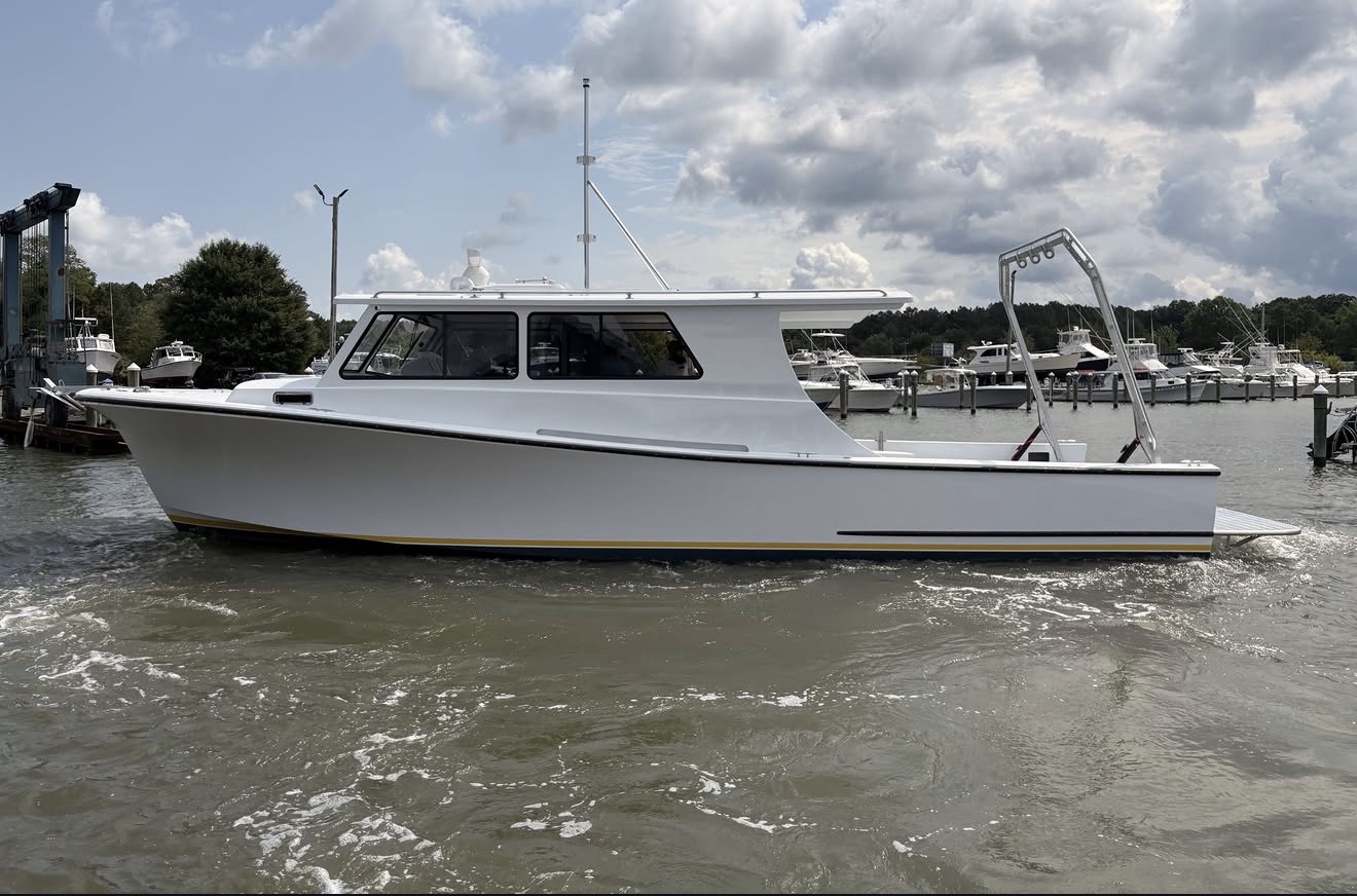 A white cabin boat is floating on the water near a marina, with other boats and trees visible in the background under a partly cloudy sky.