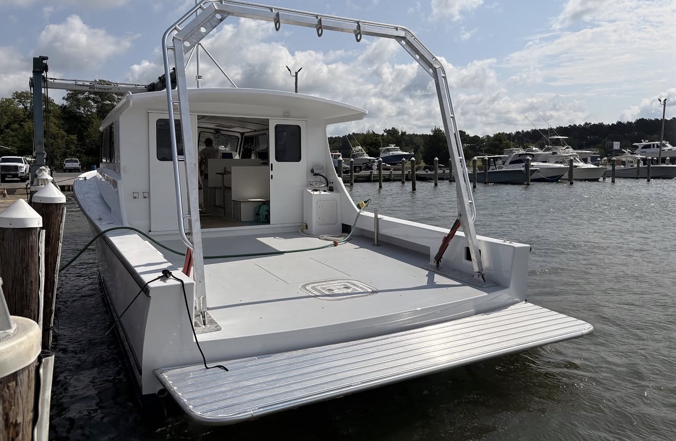 A white catamaran docked at a marina, featuring an open rear deck with a metal frame, and several boats moored in the background.