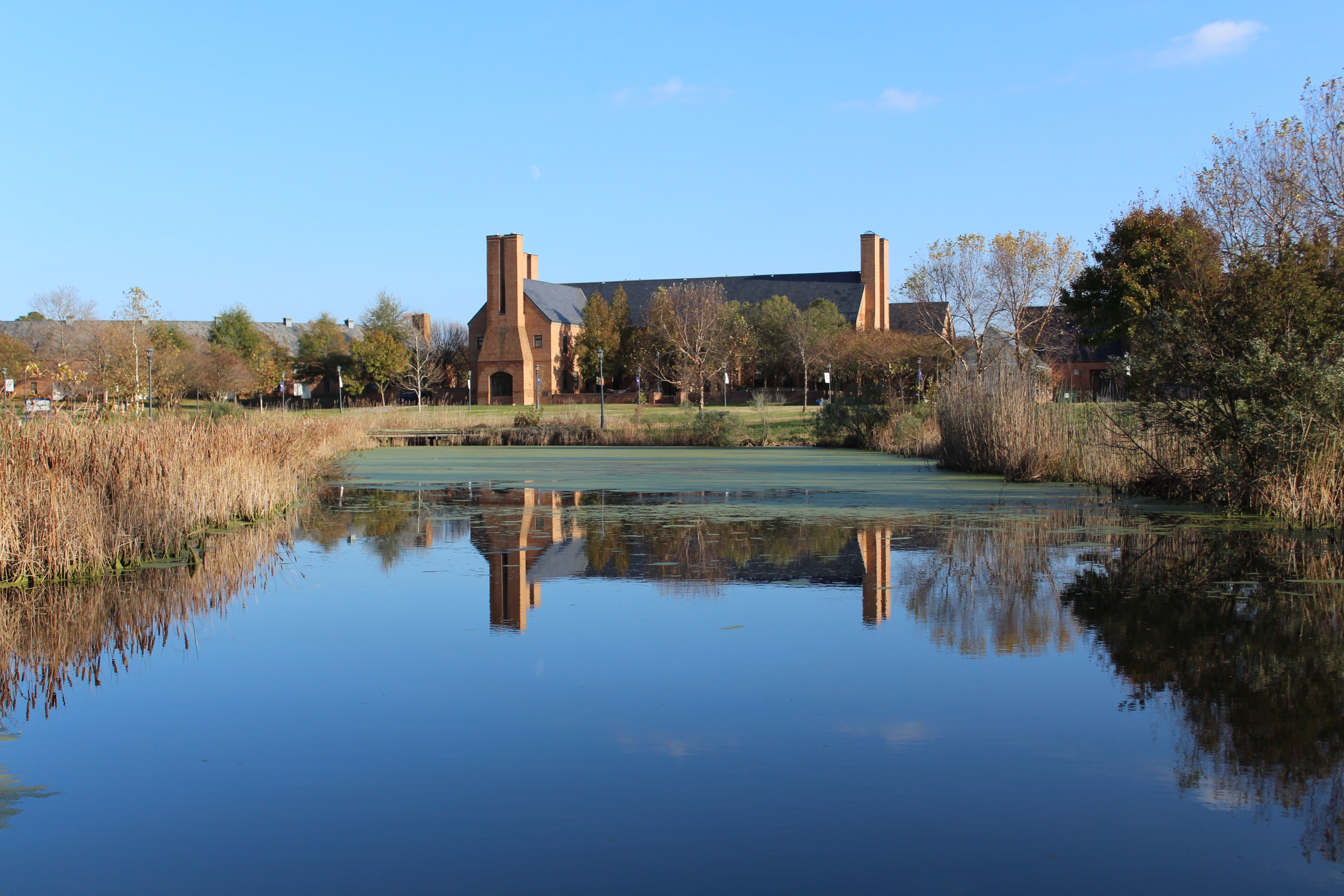 A brick building with tall chimneys is reflected in a calm pond, surrounded by reeds and trees under a clear blue sky.
