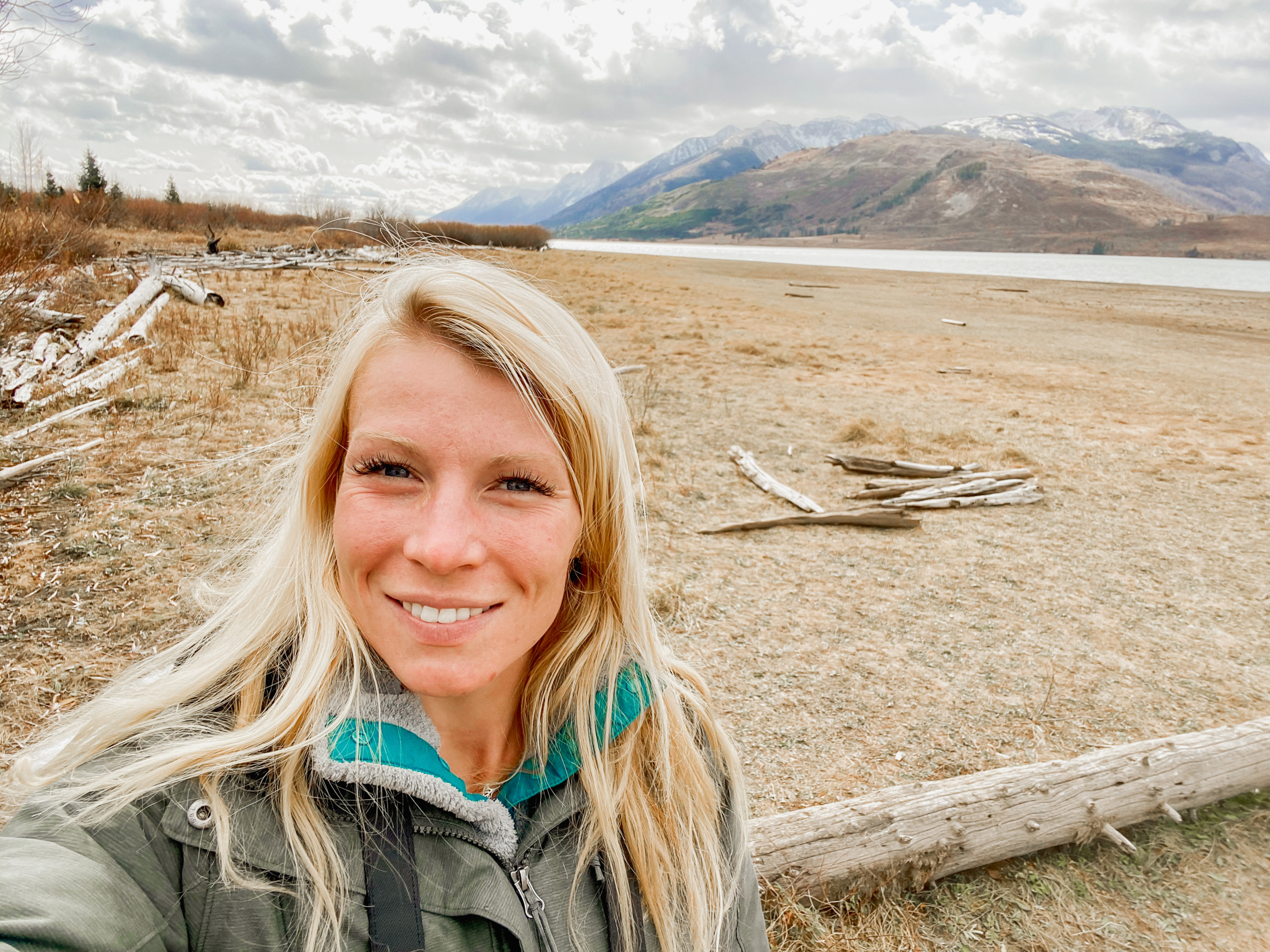 A woman with long blonde hair smiles at the camera while standing on a sandy beach with driftwood. Behind her, there are mountains with snow-capped peaks under a cloudy sky. A body of water is visible on the right side of the scene.