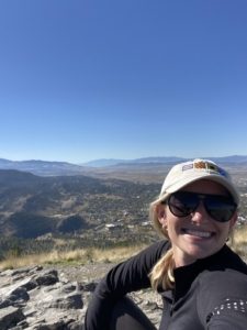 A person wearing a beige cap and sunglasses smiles while sitting outdoors. Behind them, a scenic view of rolling hills, scattered trees, and a clear blue sky.
