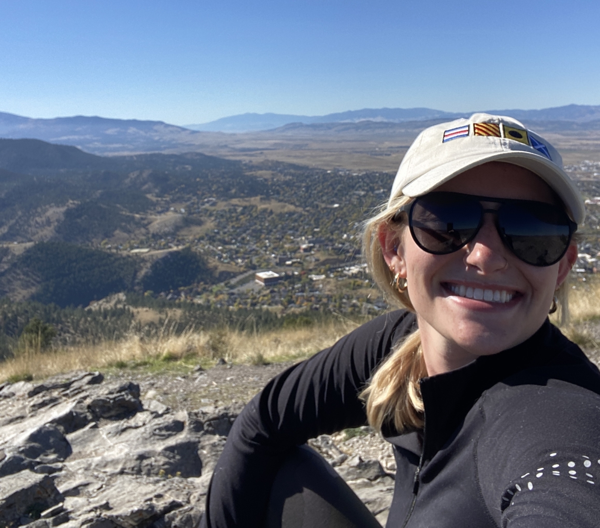 A person wearing a beige cap and sunglasses smiles while sitting outdoors. Behind them, a scenic view of rolling hills, scattered trees, and a clear blue sky.