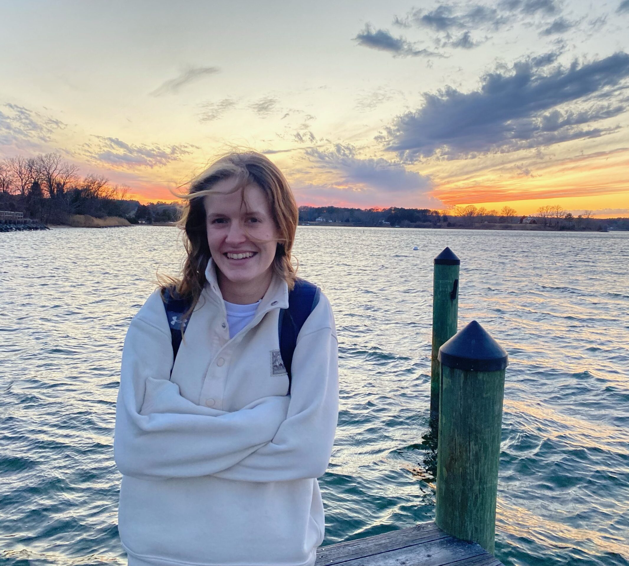 A person with light brown hair stands on a dock by a body of water at sunset, smiling with arms crossed and wearing a light-colored sweater and backpack.