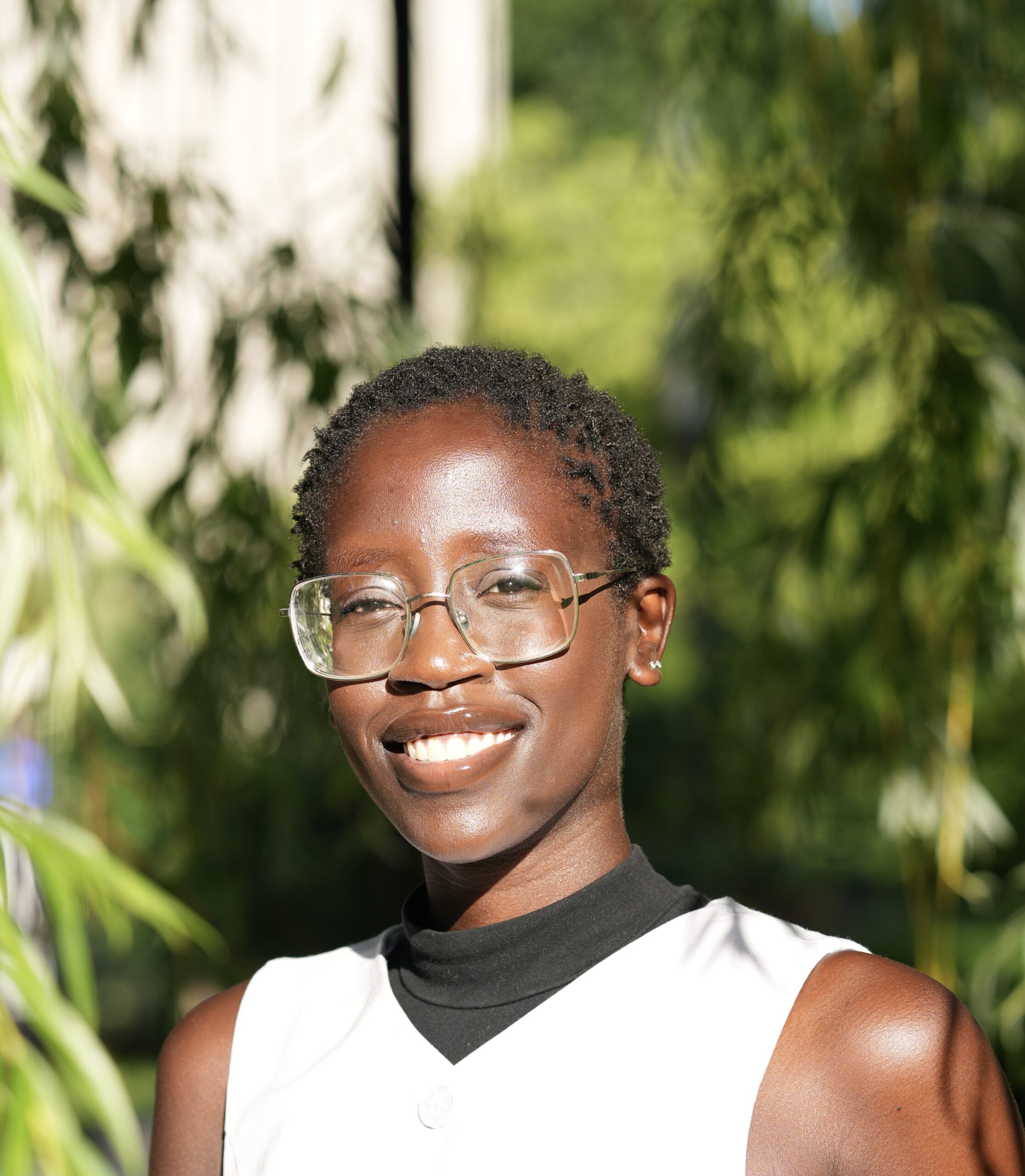 Person with short hair and glasses smiling outdoors, wearing a sleeveless white top with a black collar. Green foliage is blurred in the background.