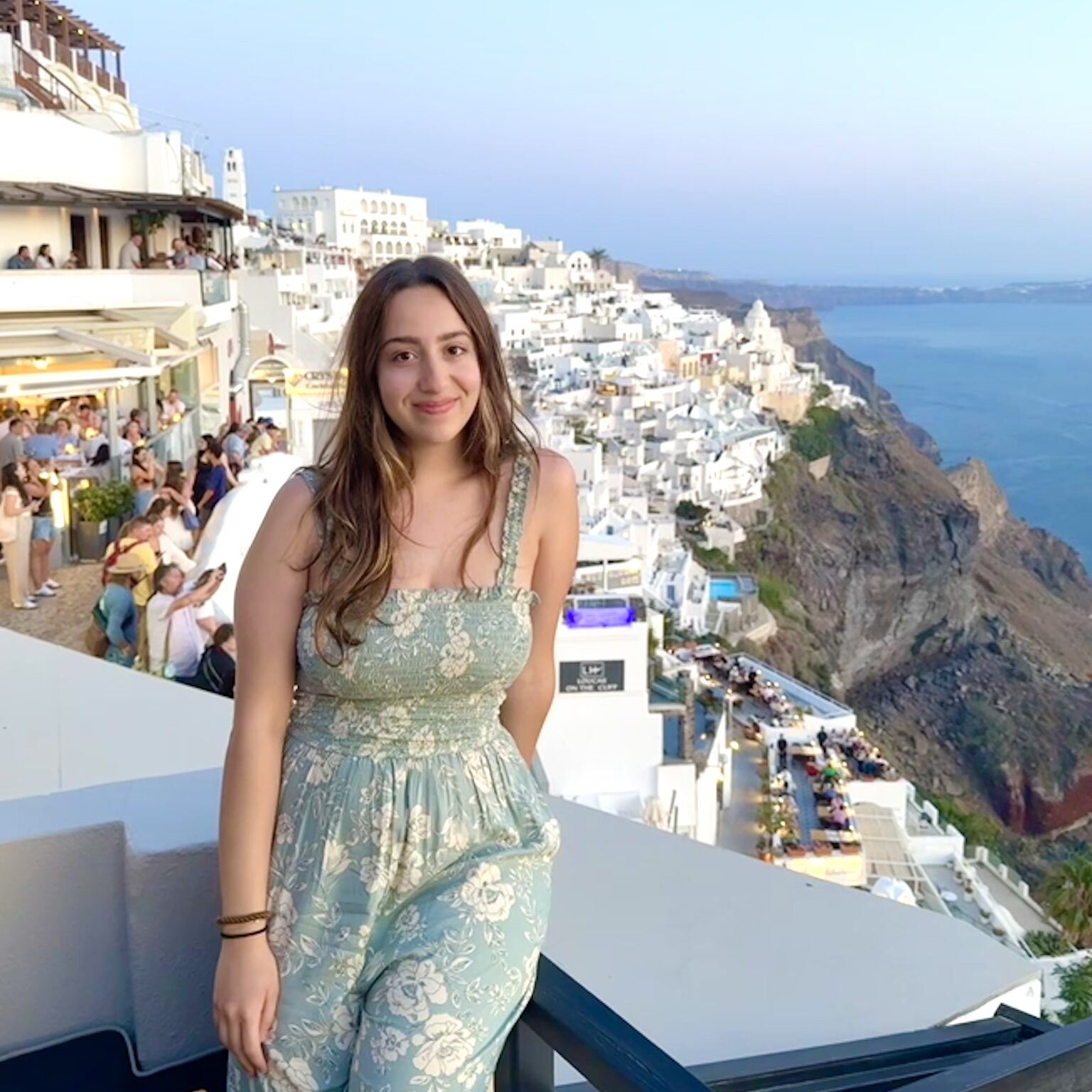 Woman in a green patterned jumpsuit poses on a terrace overlooking white buildings and the sea in Santorini, Greece at sunset.