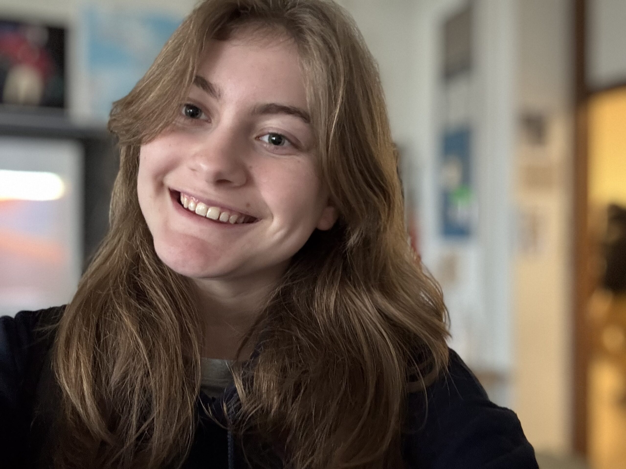 A person with long, light brown hair smiles at the camera indoors, with a blurred background featuring posters and a doorway.
