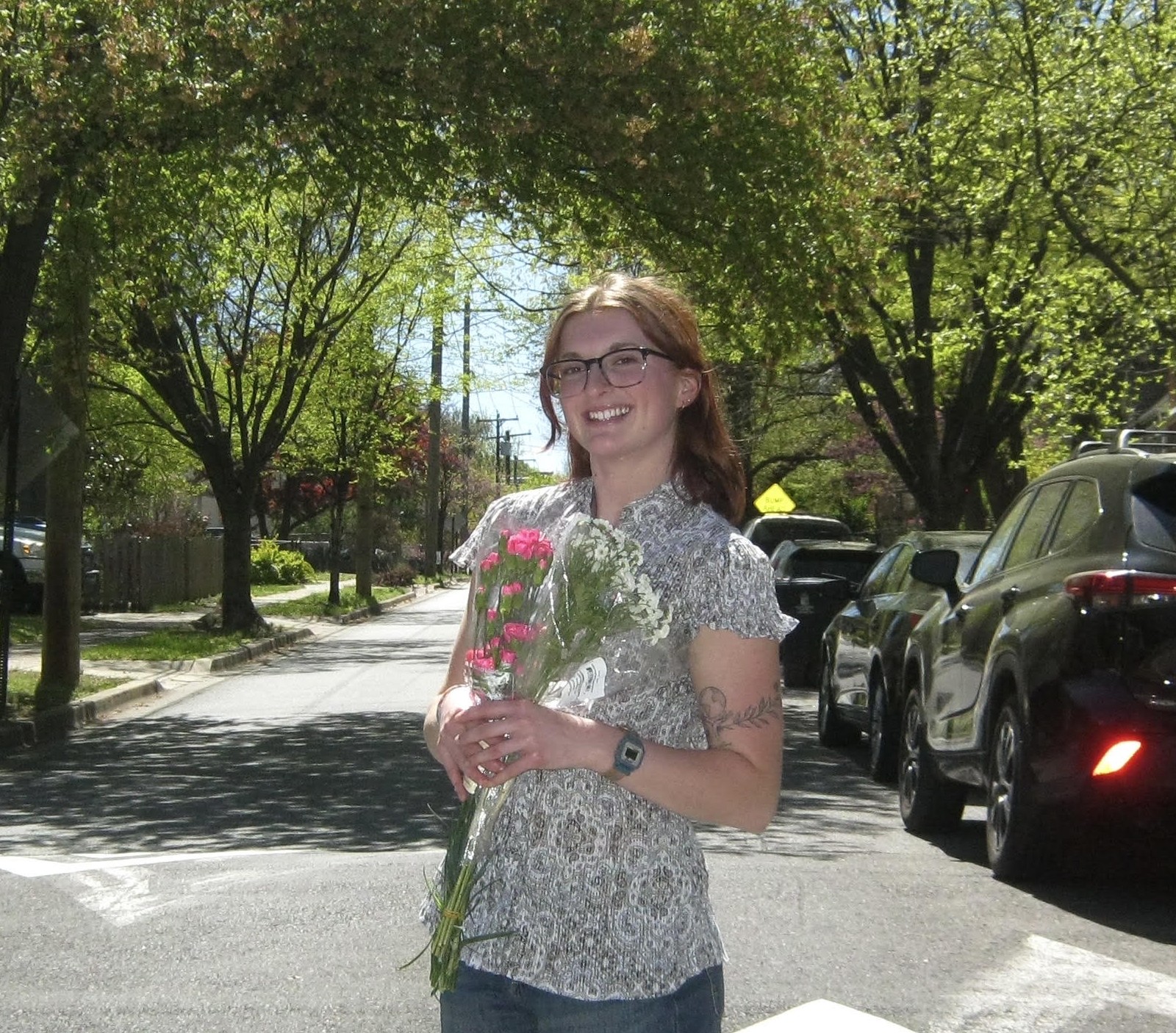 Woman standing on a sunny street holding flowers, wearing glasses, short-sleeve patterned shirt, and jeans. Trees with spring foliage and parked cars line the background.