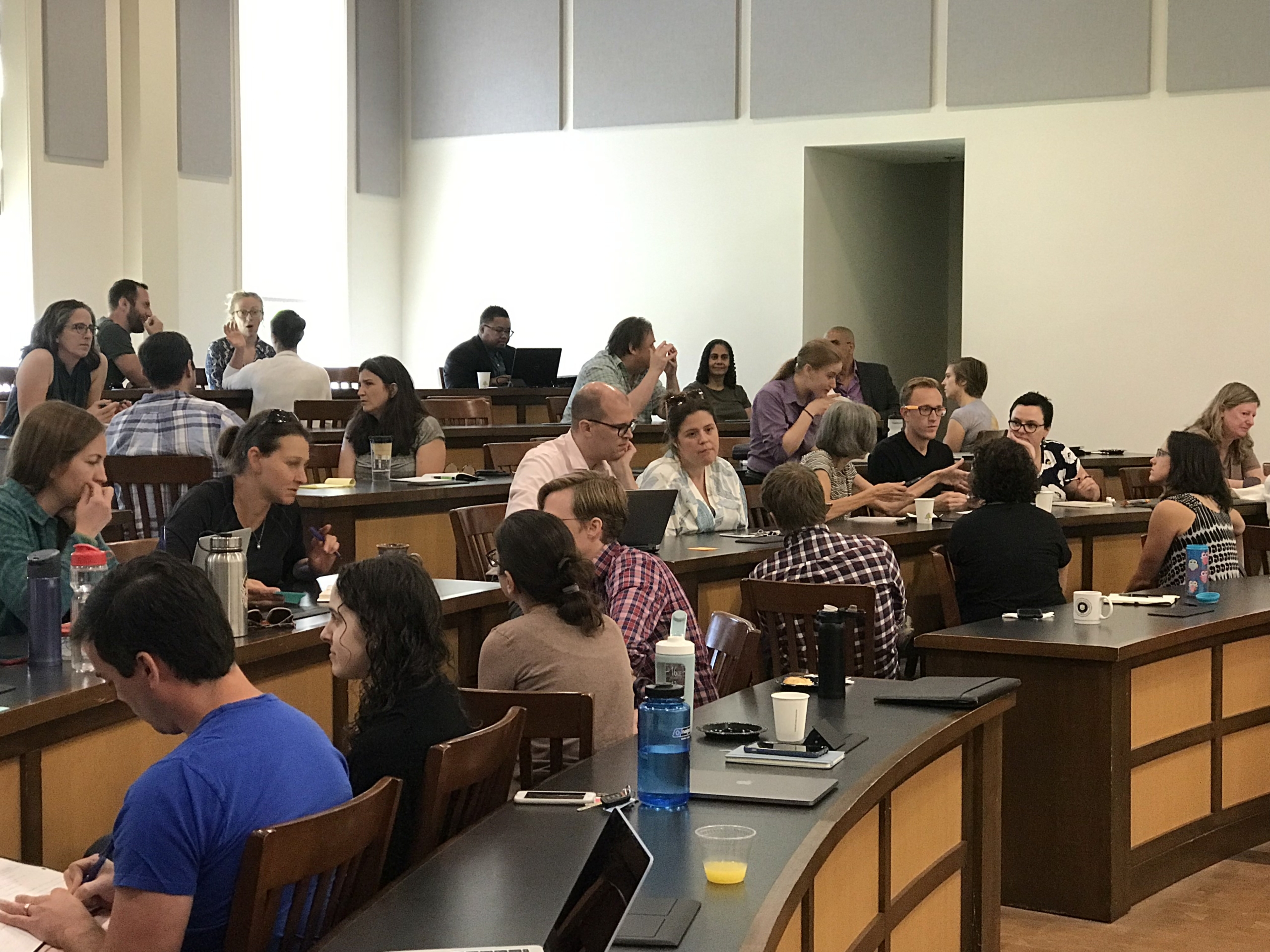 3I conference People sit in groups around tables in a classroom or seminar setting, engaged in discussion and using laptops and notebooks.