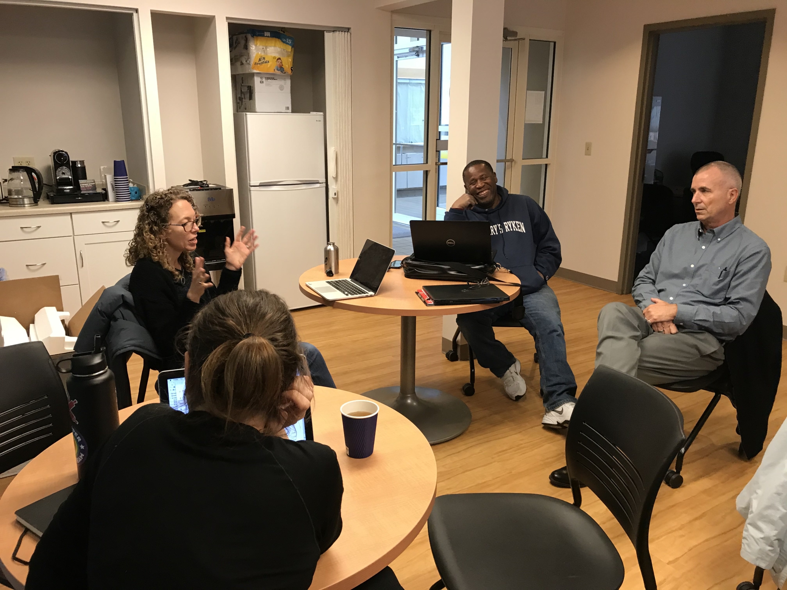 Header option 1 Four people sit around tables in an office break room, engaged in conversation; laptops, coffee cups, and notebooks are visible on the tables.