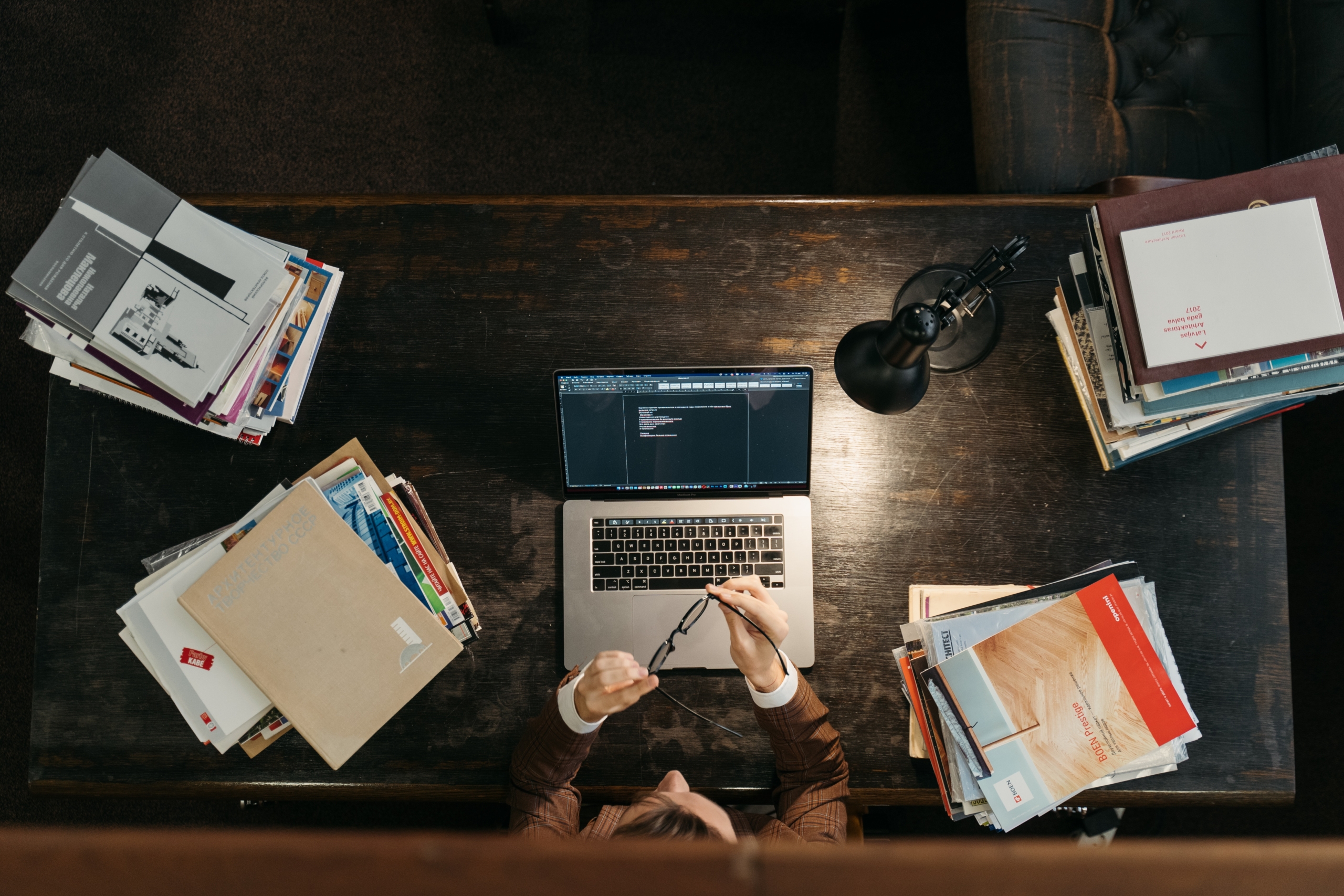 Sample STOCK PHOTO self-paced training Person sitting at a desk with an open laptop, surrounded by stacks of books and papers, holding eyeglasses, viewed from above.