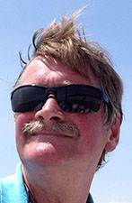 Man with a mustache wearing sunglasses and a blue shirt stands outdoors under a clear sky, wind blowing his hair—capturing the confident vibe of the St. Mary's Sailing Coaches.