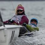Two people wearing winter gear are sailing a small boat in choppy water. Both are leaning back, suggesting they are navigating through windy conditions. The sky and water appear gray and overcast.