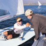 A St Mary's college of Maryland college sailor in a small boat smiles and fist-bumps a person on a nearby boat. The sailor wears a cap and sunglasses and a life vest, with a bright sky and distant cityscape in the background. Other sailboats are visible on the water.