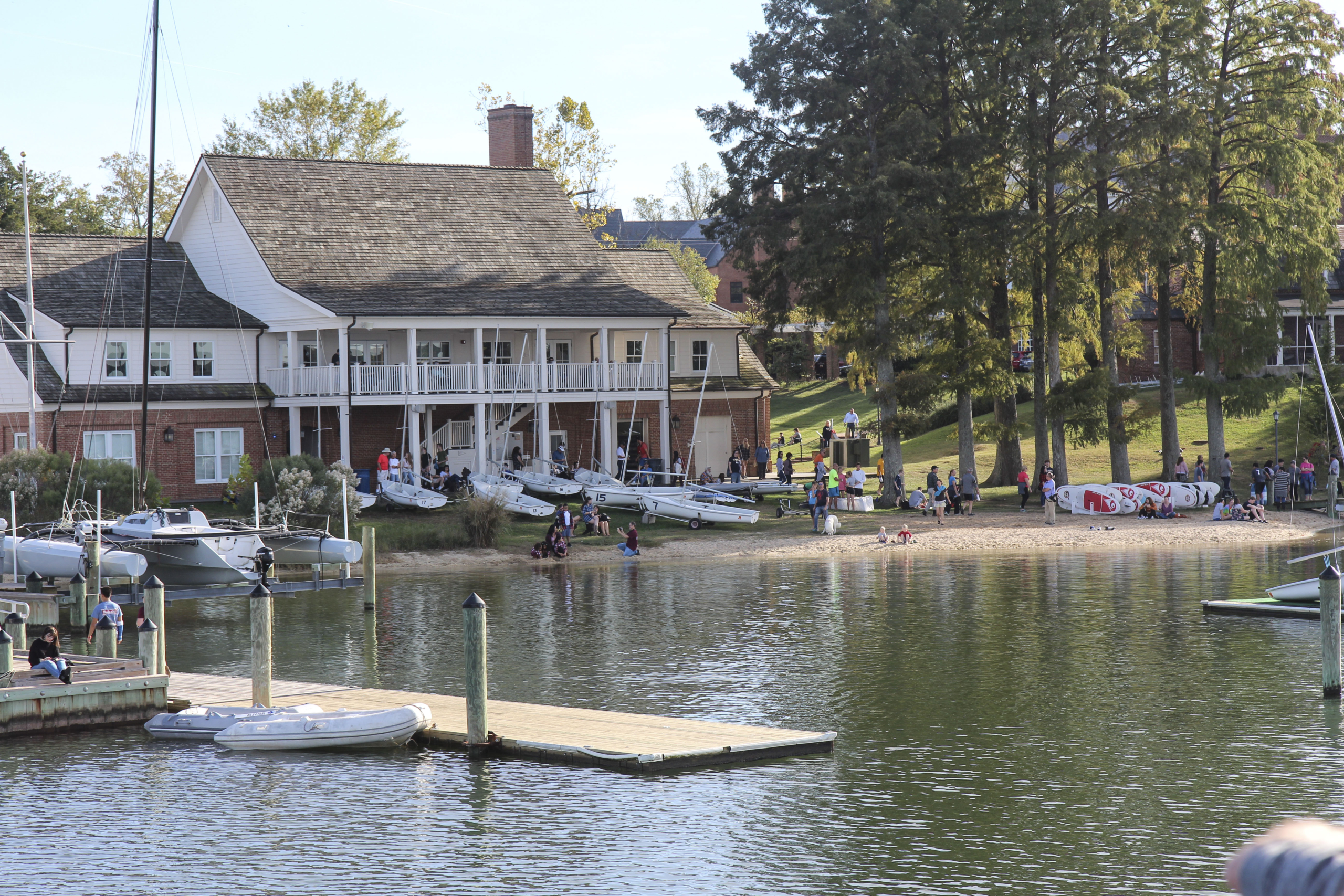 A lakeside scene with a large, two-story building featuring a porch. Several small boats and people are on the shore, with more individuals relaxing near the water's edge. Trees and a grassy lawn surround the area, creating a serene setting.