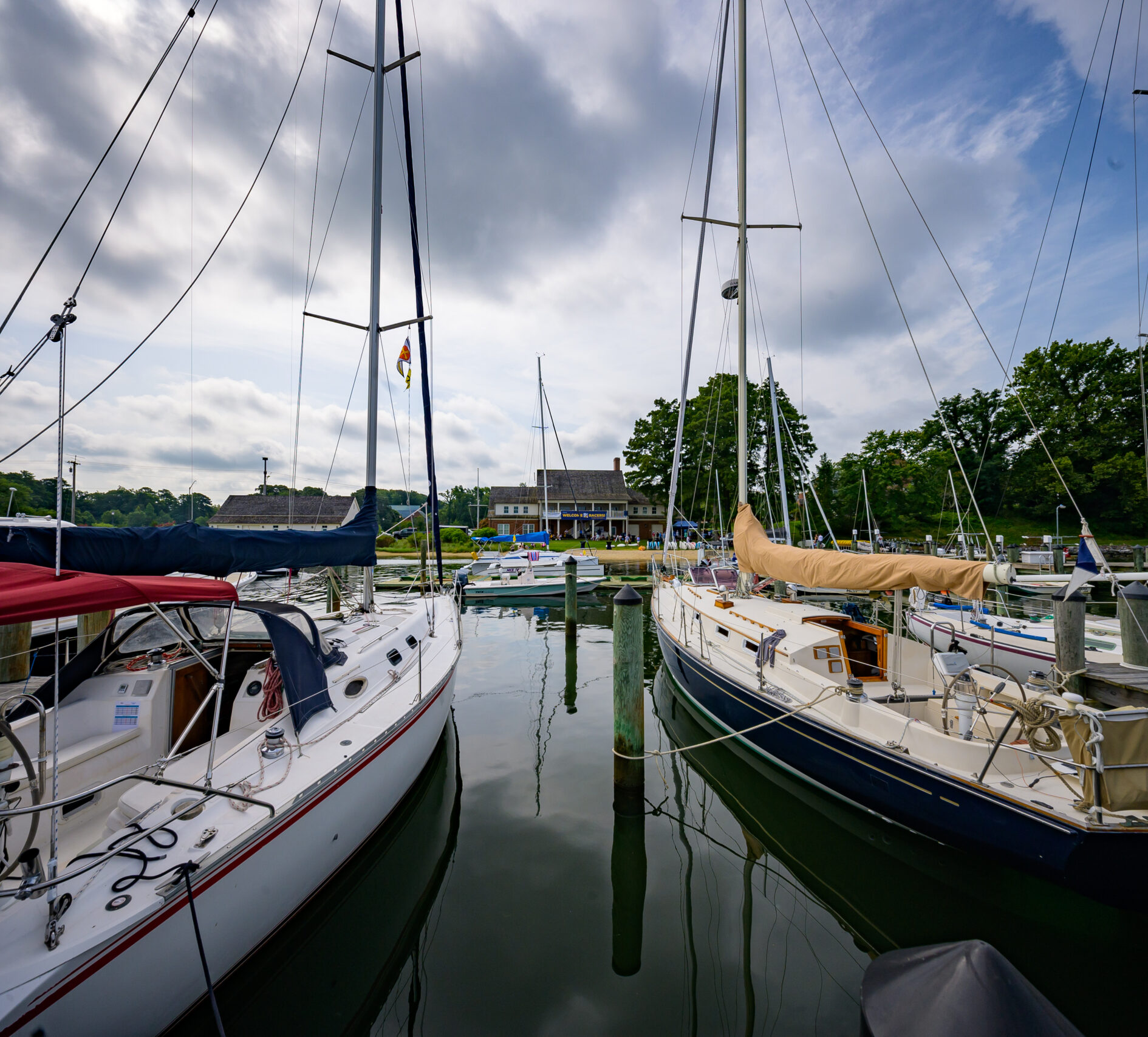 Sailboats are docked at a calm marina. The boats have various colored covers and tall masts. In the background, there's a building surrounded by trees, under a partly cloudy sky.
