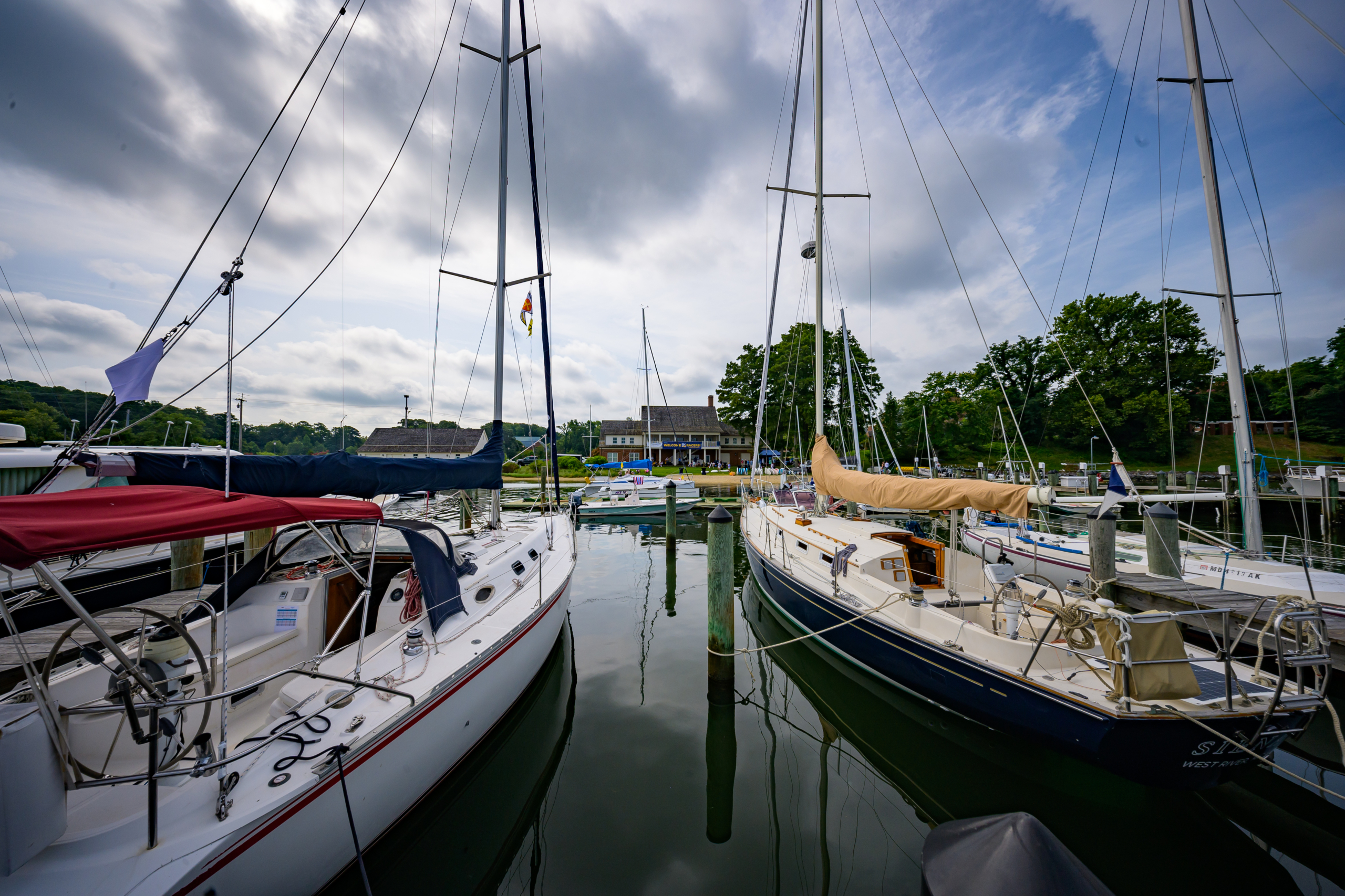 Sailboats are docked at a calm marina. The boats have various colored covers and tall masts. In the background, there's a building surrounded by trees, under a partly cloudy sky.