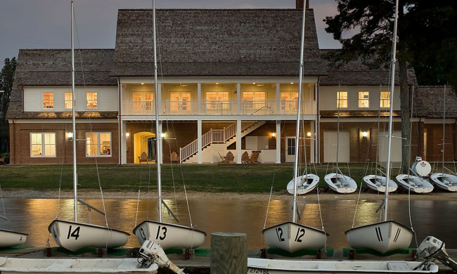 A large building with warm interior lights sits by the water. In front, a row of small sailboats with numbered sails are docked. The scene is calm, with a soft evening glow.