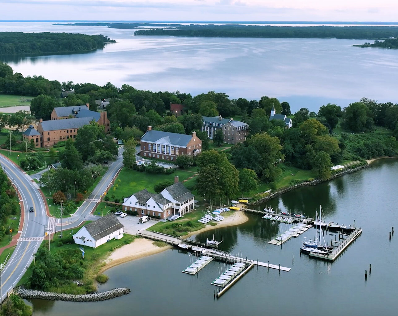 Aerial view of a smcm waterfront with boats docked at a marina, several buildings, a curving road, and surrounding trees by a large body of water.