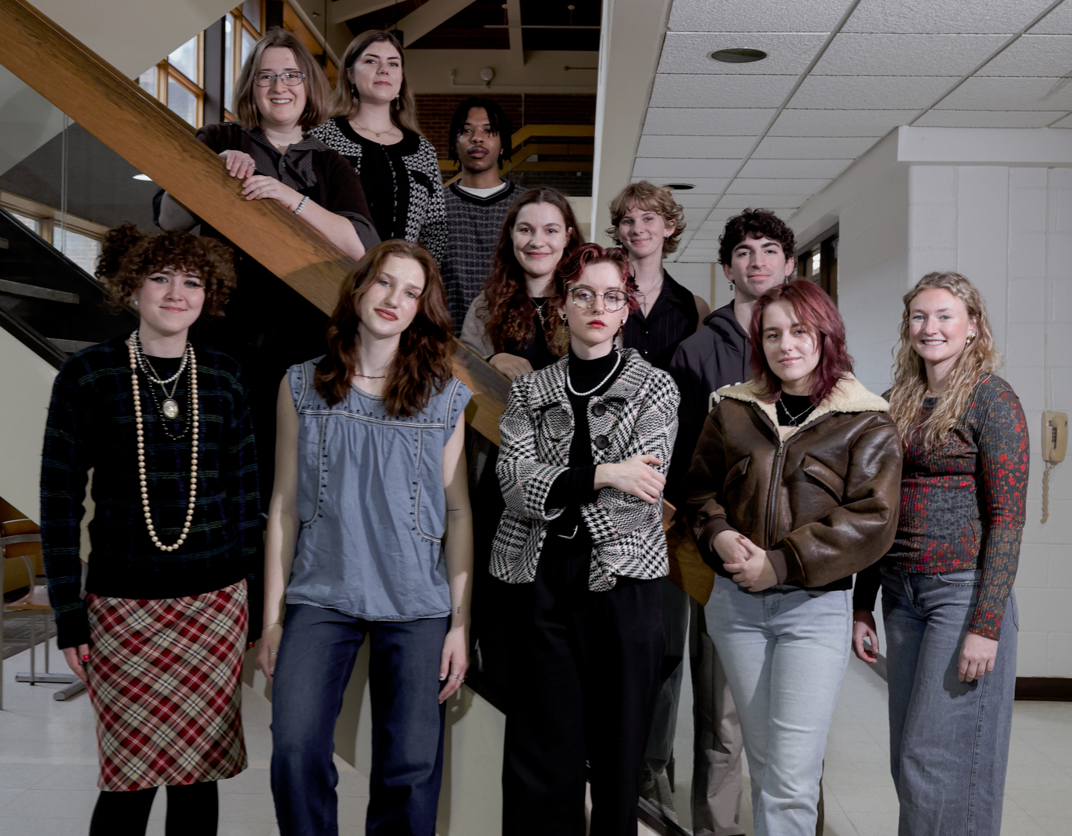 A group of eleven young adults pose for a photo on and around a staircase inside a building, with neutral expressions and casual to semi-formal attire.