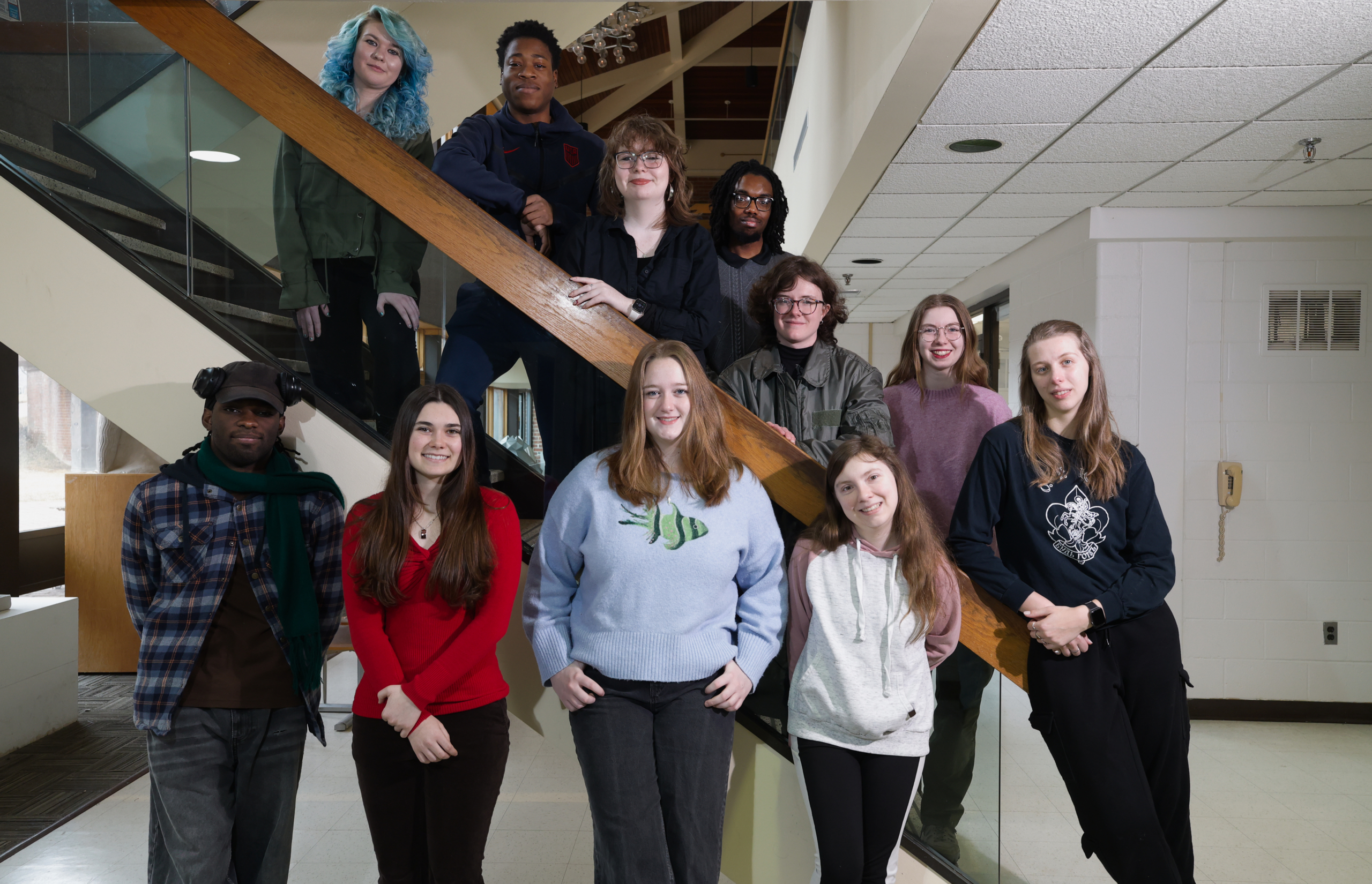 Ten people pose on and around a staircase inside a building with white walls and a tiled ceiling. Some stand on the steps, others on the floor, all facing the camera and smiling.