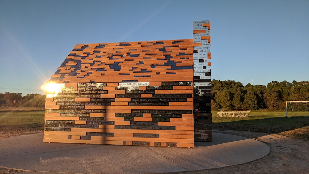 A small wooden structure composed of alternating wood and metal slats stands on a concrete pad in an open grassy field under a clear sky.
